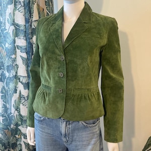 May include: A green suede blazer with a notched lapel, three-button closure, and a small pocket. The blazer has a fitted silhouette and a slightly flared hem. The jacket is displayed on a mannequin.