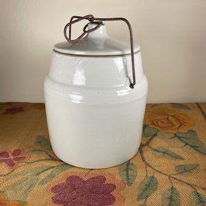 Puede incluir: Una vasija de cerámica cilíndrica de color blanco roto con un acabado esmaltado liso y una parte superior ligeramente cónica. El recipiente incluye una tapa ajustada con un pomo central y un asa de alambre metálico oxidado.