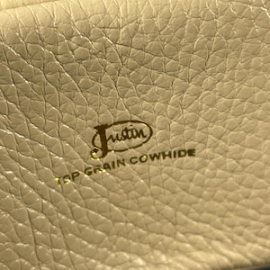 May include: Close-up of a cream-colored leather surface with a textured grain. The gold-colored text reads "Justin TOP GRAIN COWHIDE" in a stylized font, indicating the material and brand.