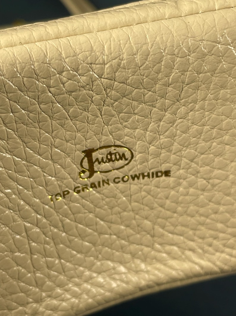 May include: Close-up of a cream-colored leather surface with a textured grain. The gold-colored text reads "Justin TOP GRAIN COWHIDE" in a stylized font, indicating the material and brand.