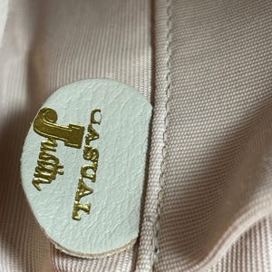 May include: Close-up of a light green leather tag with the gold text "Justin Casual". The tag is circular and attached to a light pink fabric. The fabric has a woven texture.