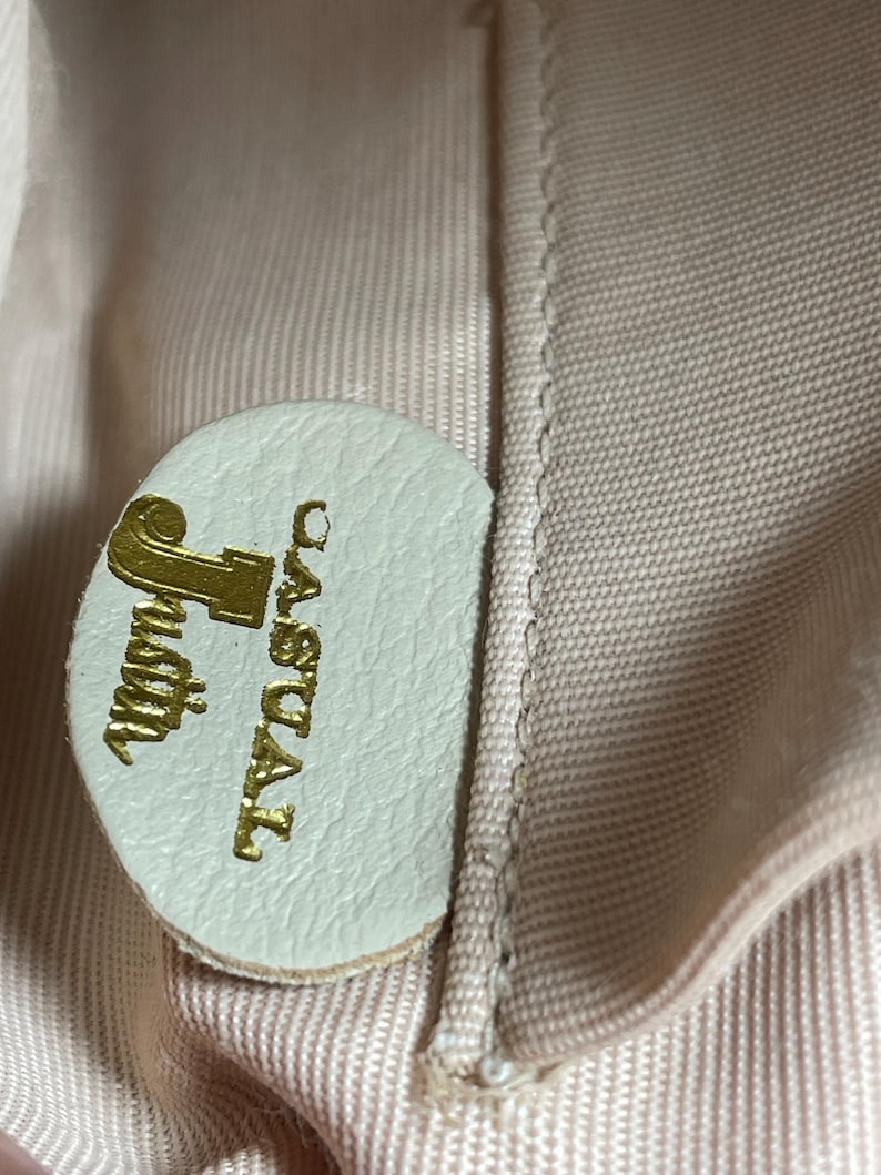 May include: Close-up of a light green leather tag with the gold text "Justin Casual". The tag is circular and attached to a light pink fabric. The fabric has a woven texture.