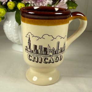 Puede incluir: Taza de cerámica color crema con borde y asa marrones. La taza presenta una ilustración del horizonte con la palabra "CHICAGO" en negro. La taza tiene una base de pedestal. Flores en el fondo.