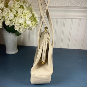 May include: Cream-colored vintage handbag with a structured design. The bag features a clasp closure with gold-tone hardware and dual top handles. The purse is made of a textured material and has a rectangular base.