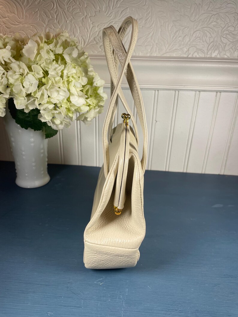 May include: Cream-colored vintage handbag with a structured design. The bag features a clasp closure with gold-tone hardware and dual top handles. The purse is made of a textured material and has a rectangular base.
