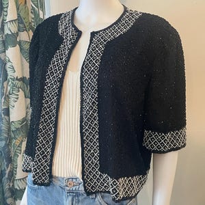 May include: Black beaded jacket with white geometric trim. The short-sleeved jacket features a round neckline and is displayed on a mannequin. The jacket is paired with a white top and blue jeans.
