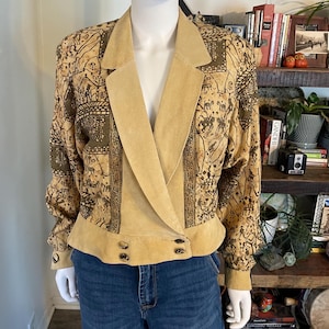May include: A tan suede blazer featuring a dark brown printed pattern of stylised figures and animals. The jacket has a wide lapel, long sleeves with buttoned cuffs, and a cropped hemline with two sets of buttons at the front waist area.