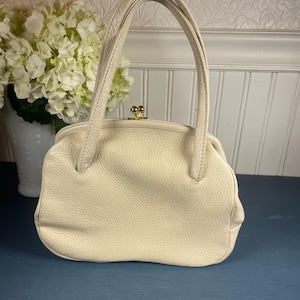 May include: Cream-colored leather handbag with a rounded shape and short handles. The bag features a textured surface and a gold-colored clasp. A vintage-style purse, perfect for carrying essentials.