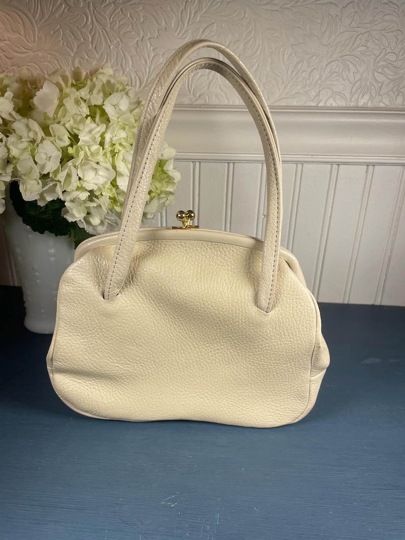 May include: Cream-colored leather handbag with a rounded shape and short handles. The bag features a textured surface and a gold-colored clasp. A vintage-style purse, perfect for carrying essentials.