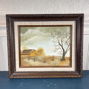 May include: Framed landscape painting depicting a rural scene with a barn, fence, and leafless tree under a cloudy sky. The painting is in a dark brown ornate frame, adding a vintage aesthetic. The artwork evokes a sense of tranquility.