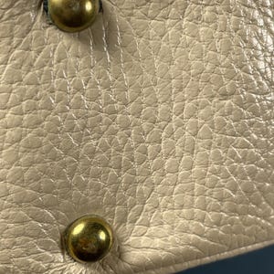 May include: Close-up of a cream-colored leather surface with a textured grain. Two brass-colored, rounded studs are visible, one at the top and one at the bottom. The leather appears to be part of a larger item, possibly a bag or accessory.
