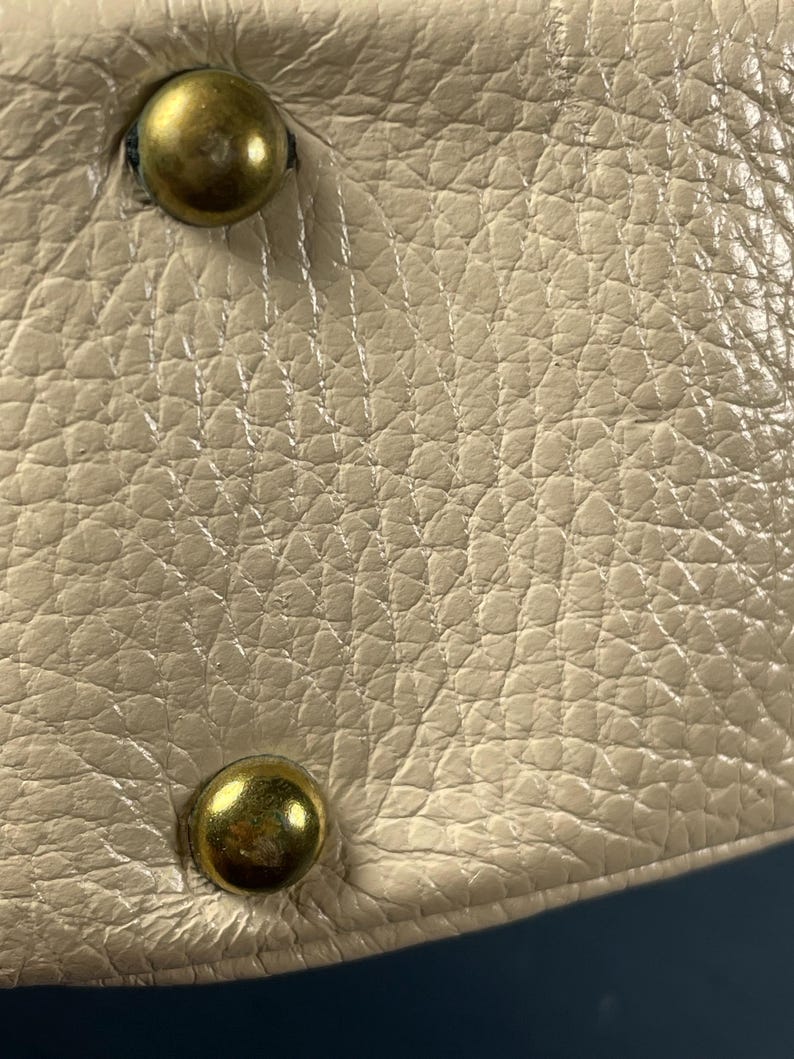 May include: Close-up of a cream-colored leather surface with a textured grain. Two brass-colored, rounded studs are visible, one at the top and one at the bottom. The leather appears to be part of a larger item, possibly a bag or accessory.