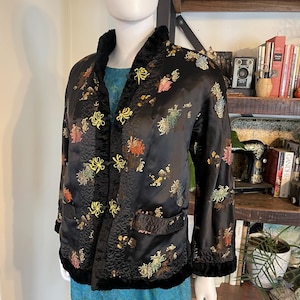 Puede incluir: Chaqueta de satén negro con bordados florales en tonos amarillos, rojos y verdes. La chaqueta presenta un diseño acolchado, ribete de terciopelo negro y cierres de rana. Las mangas son largas y la chaqueta tiene dos bolsillos.