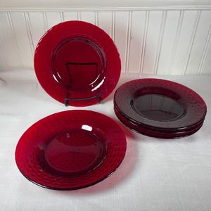 Puede incluir: Un juego de cuatro platos de vidrio rojo con una superficie texturizada y con hoyuelos. Un plato se muestra en un soporte de metal negro, mientras que los otros están apilados. Los platos son de color rojo intenso y miden unos 25 cm de diámetro.