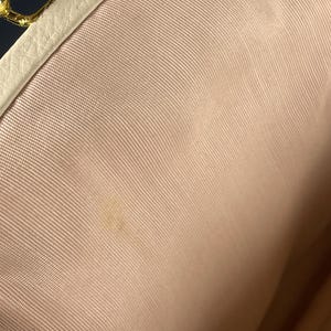 May include: Interior view of a light pink fabric purse. The purse has a cream-colored leather trim and a gold-colored clasp. The fabric has a subtle texture and a small stain is visible.