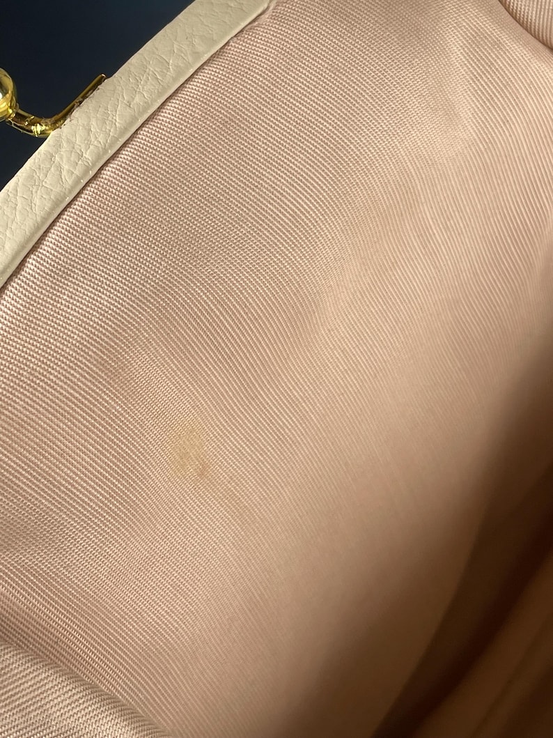 May include: Interior view of a light pink fabric purse. The purse has a cream-colored leather trim and a gold-colored clasp. The fabric has a subtle texture and a small stain is visible.