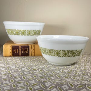 May include: Two white milk glass mixing bowls featuring a repeating olive green geometric floral pattern around the exterior. The bowls are positioned on a yellow book titled The Good Housekeeping Cook Book and a patterned fabric surface.