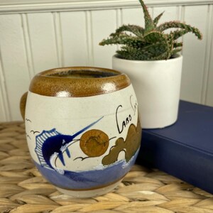 May include: A round ceramic mug with a speckled cream finish and a brown rim. The mug features a hand-painted blue marlin fish, a brown sun, and rock formations above blue waves. The text Cabo San is written in black script on the side.