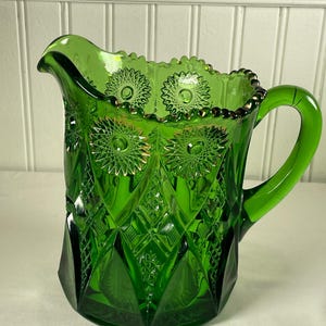 Puede incluir: Una jarra de vidrio verde con un patrón decorativo. La jarra tiene un asa y un pico. El vidrio tiene un diseño de diamantes y estrellas. El borde está adornado con un borde de cuentas de color dorado. La jarra mide aproximadamente 20 cm de alto.