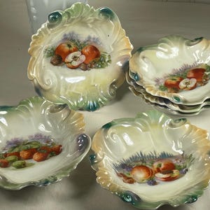 May include: A collection of decorative porcelain dishes with scalloped edges. The dishes feature hand-painted fruit designs, including apples, strawberries, and peaches, with green, yellow, and blue accents. The set includes individual dishes and a stack of smaller ones.