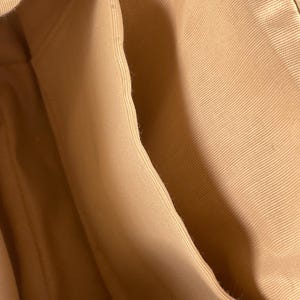 May include: Close-up of the interior of a beige handbag, showcasing the fabric lining and pockets. The light tan fabric has a subtle texture, with visible seams and folds. The bag's design includes multiple compartments.