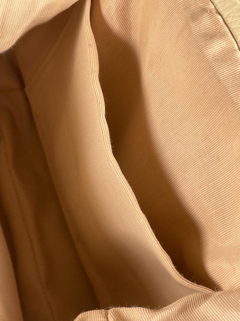May include: Close-up of the interior of a beige handbag, showcasing the fabric lining and pockets. The light tan fabric has a subtle texture, with visible seams and folds. The bag's design includes multiple compartments.