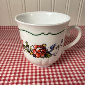 May include: White ceramic teacup with a floral design featuring red, blue, and yellow flowers. The cup has a green trim and a curved handle. The cup is sitting on a red and white checkered tablecloth.
