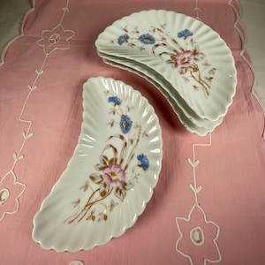 May include: White, fan-shaped porcelain dishes with a ruffled edge. Each dish is decorated with a floral design featuring pink and blue flowers with brown stems and leaves. The dishes are stacked on a pink tablecloth.