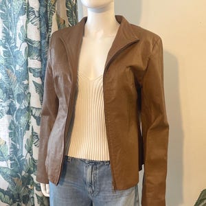 May include: A brown jacket with a subtle perforated texture, featuring a full-length front zipper and a stand-up collar. The jacket is worn over a cream-colored ribbed tank top and light blue denim jeans, displayed on a mannequin torso.