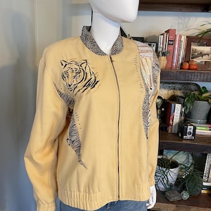 May include: A light yellow jacket with a tiger design on the front. The jacket has a black and white animal print collar and a zipper closure. The jacket is on a mannequin.