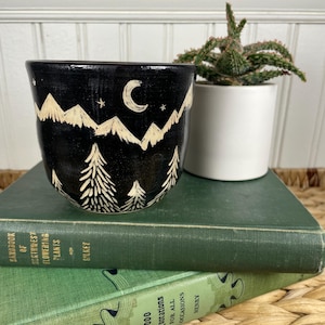 May include: A black ceramic mug featuring a hand-carved cream-colored design of a mountain range, pine trees, a crescent moon, and stars. The mug sits atop two stacked green books, one titled Handbook of Northwest Flowering Plants.