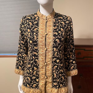 May include: A black jacket featuring a mandarin collar and three-quarter length sleeves. The garment is adorned with an intricate gold and orange paisley embroidery pattern throughout, and it fastens down the front with four round fabric-covered buttons.