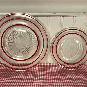 Peut inclure: Deux assiettes en verre transparent avec des anneaux rouges concentriques. La plus grande assiette a un motif en étoile au centre. Les assiettes sont sur une nappe à carreaux rouges et blancs.