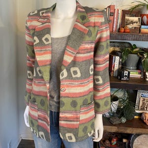Può includere: Un blazer con un motivo multicolore a righe orizzontali, cerchi scuri e forme quadrate chiare. Il capo presenta maniche lunghe, un bavero dentellato e due tasche frontali applicate, indossato sopra un top grigio e un paio di jeans blu.