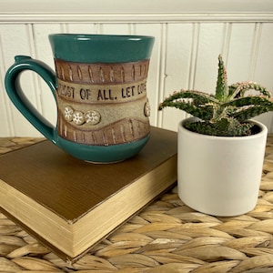 May include: A teal ceramic mug with a brown textured band featuring the embossed text "MOST OF ALL, LET LOVE" and small flower accents. Beside it sits a green succulent plant in a plain white cylindrical ceramic pot on a brown book.