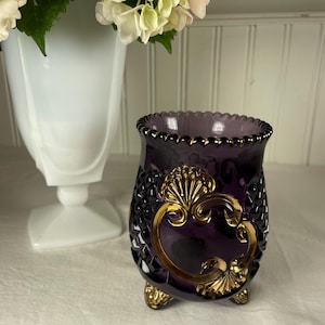 Puede incluir: Un jarrón decorativo de cristal color amatista con un diseño dorado ornamentado. El jarrón tiene un patrón de diamantes y se apoya en tres pequeños pies. Un jarrón blanco y flores blancas en el fondo.