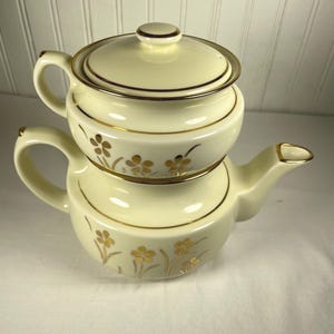 May include: Cream-coloured ceramic teapot with a matching lid and a separate creamer. The teapot and creamer are adorned with gold trim and floral designs. The teapot has a curved spout and a handle. The creamer sits atop the teapot.