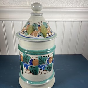 May include: A ceramic jar with a lid, featuring a hand-painted floral design in shades of blue, green, and brown. The jar has a cylindrical body with a turquoise band and a matching lid with a knob. The background is a white wall.