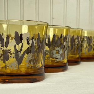 Könnte beinhalten: Vier bernsteinfarbene Trinkgläser mit einem botanischen Design. Die Gläser haben einen dunkelbraunen Boden und sind mit einem Muster aus braunen, grauen und weißen Pflanzen verziert. Die Gläser sind in einer Reihe angeordnet.