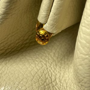 May include: Close-up of a cream-colored leather handbag with a gold-colored clasp. The clasp is round with a decorative design. The leather has a textured, pebbled appearance.
