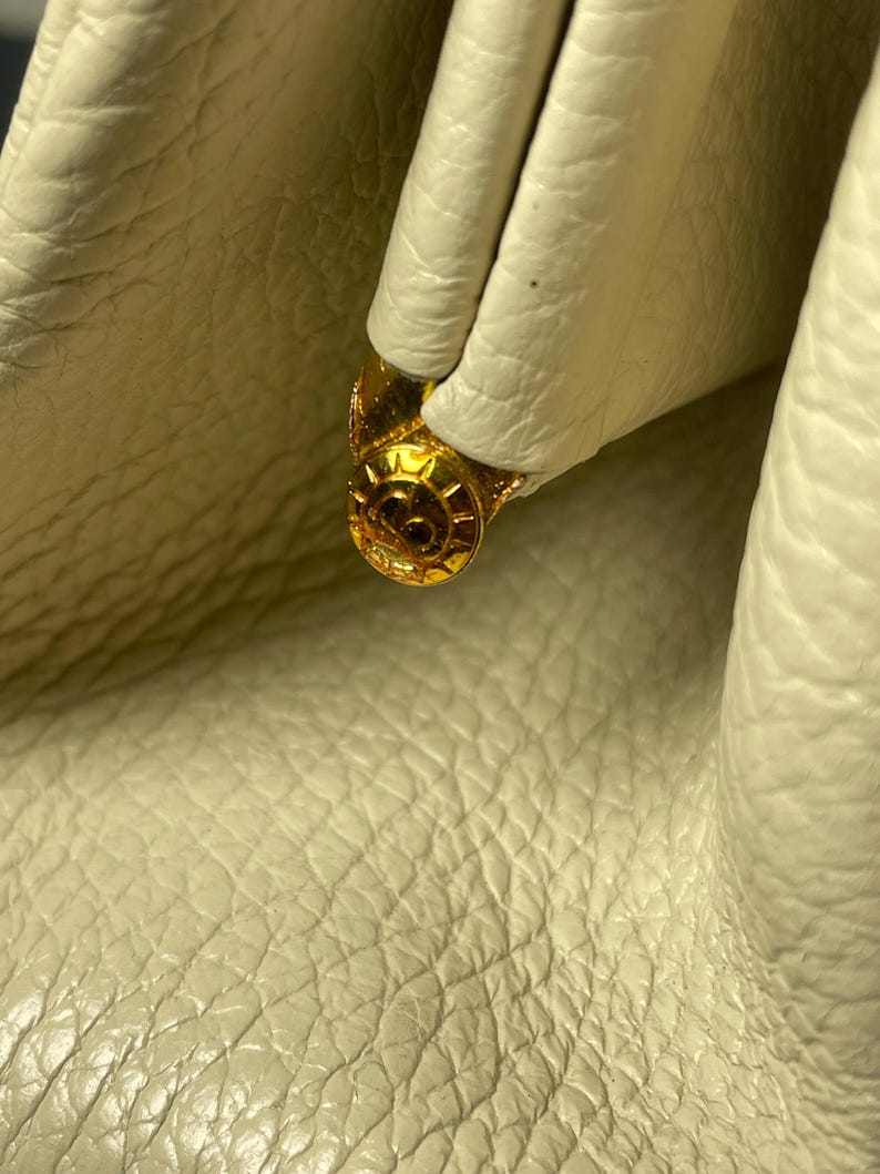 May include: Close-up of a cream-colored leather handbag with a gold-colored clasp. The clasp is round with a decorative design. The leather has a textured, pebbled appearance.