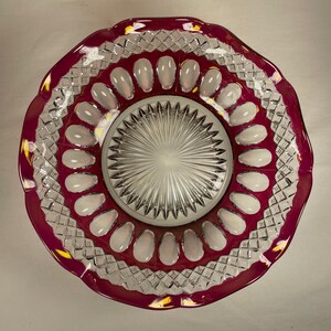 May include: A decorative glass bowl with a ruby red rim and a clear, textured center. The bowl features a scalloped edge and a pattern of clear, oval-shaped accents. The center has a starburst design.