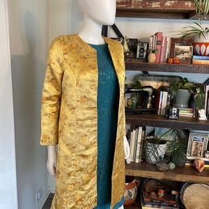 以下が含まれることがあります： マネキンに展示されている、花柄のゴールドカラーの長袖コート。 コートは開いており、下にティールカラーのドレスが見えます。 コートは丸いネックラインと膝丈の裾です。 背景には本棚があります。