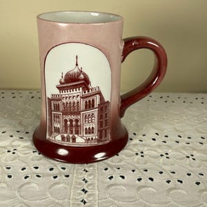 May include: A ceramic mug with a flared base, featuring a gradient color scheme that transitions from a light pink at the top to a deep maroon at the bottom. A white arched panel on the front displays a detailed dark red illustration of a building.