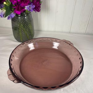 Puede incluir: Un molde para tarta Pyrex morado vintage, redondo, con borde festoneado y dos asas. El plato es de vidrio transparente. Un jarrón de flores moradas está en el fondo. El plato mide aproximadamente 25 cm de diámetro.