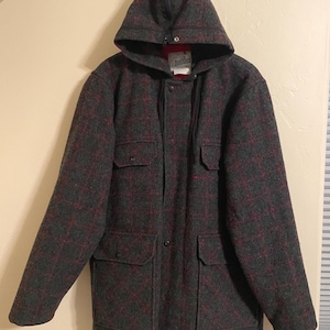 May include: A charcoal gray hooded jacket featuring a dark red plaid pattern. The jacket includes a front zipper closure, a snap-button placket, two chest pockets with flaps, and two lower pockets with flaps. A white label with black text is visible.