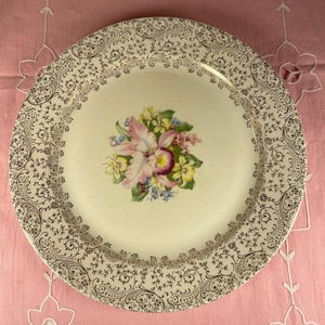 Könnte beinhalten: Ein cremefarbener Vintage-Teller mit einem Blumenzentrum mit rosa, gelben und blauen Blumen. Der Teller hat einen gold-schwarzen Filigranrand. Der Teller ist rund und liegt auf einer rosa Oberfläche.