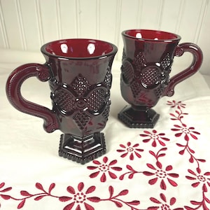 Op de afbeelding: Twee robijnrode glazen mokken met sierlijke, getextureerde ontwerpen en handvatten. De mokken staan op een wit oppervlak met een rode bloemen geborduurde rand. De mokken hebben een voetstuk en zijn waarschijnlijk vintage of antiek.