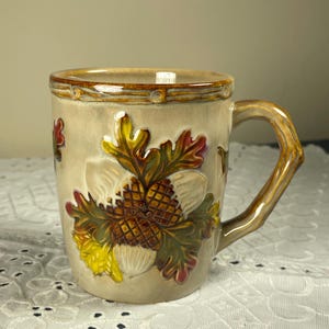 May include: A ceramic mug with a beige base color featuring a raised, three-dimensional relief design of brown acorns and multi-colored oak leaves in shades of green, yellow, and red. The mug has a textured handle shaped like a tree branch.
