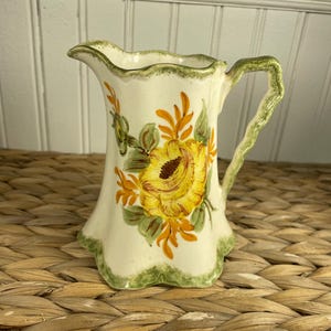 Può includere: Una brocca in ceramica color crema con un fiore giallo dipinto a mano e foglie arancioni e verdi. La brocca presenta un bordo e una base smerlati, con un bordo dipinto in verde testurizzato e un manico verde abbinato dal design ondulato.